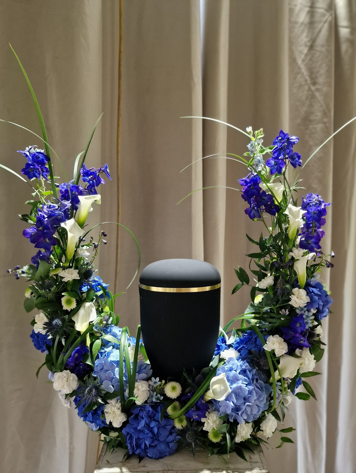 Schwarze Urne in einem offenen Blumenbogen aus blauem Rittersporn, Hortensien und weißen Callas