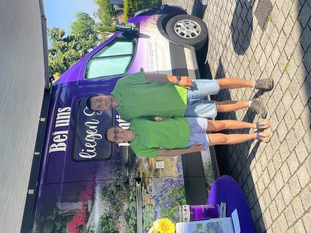 Zwei Mitarbeitende in grünen Firmenpoloshirts vor dem Aktionsfahrzeug zur Dauergrabpflege