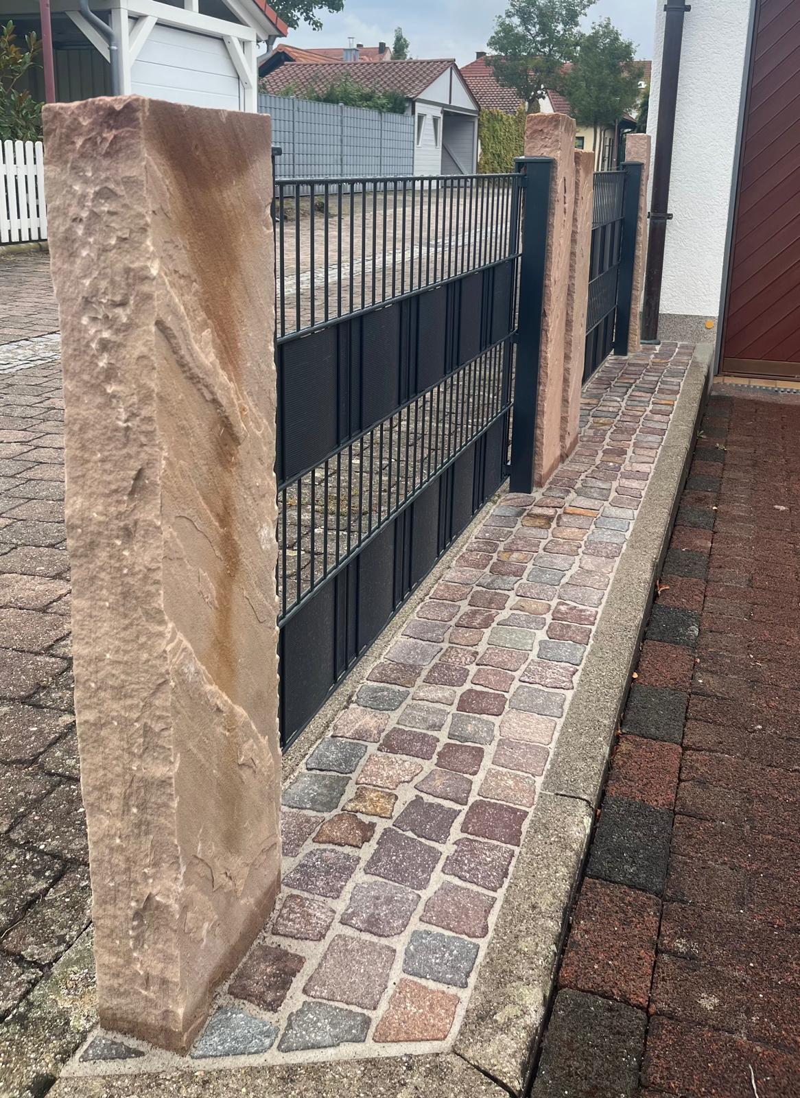 Anthrazitfarbener Zaun zwischen Sandsteinstelen mit Natursteinpflaster-Streifen