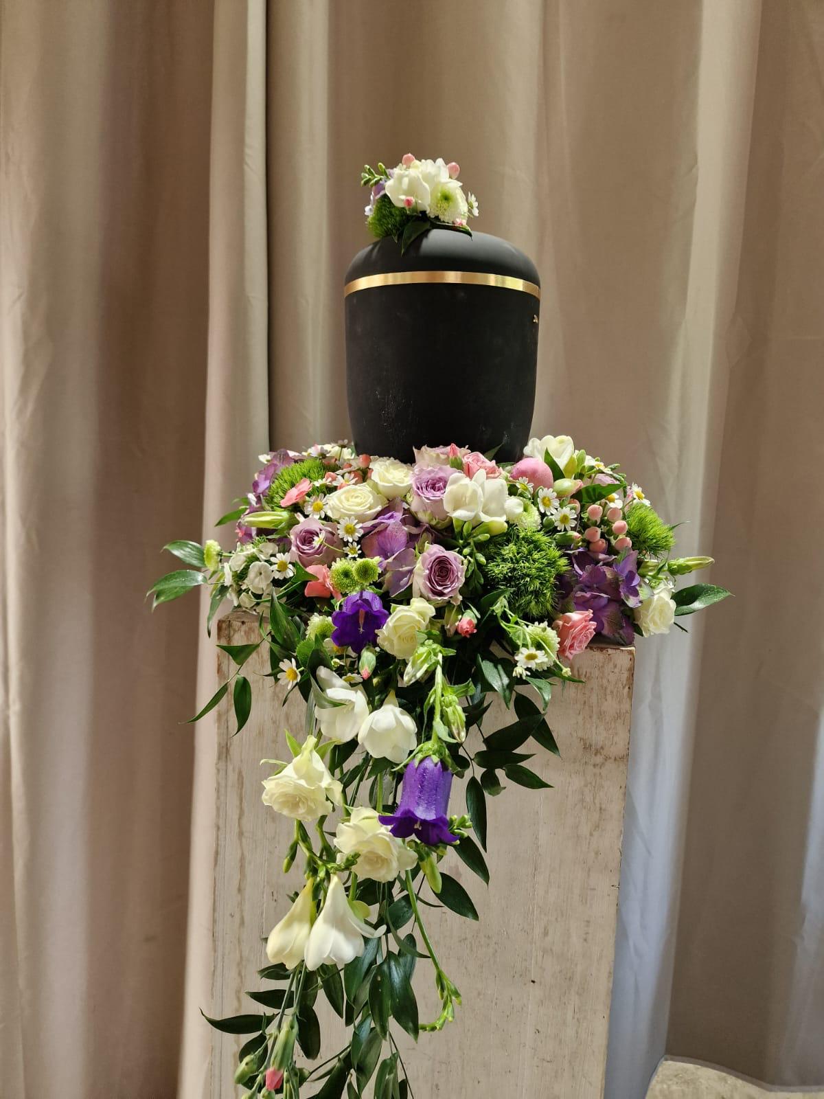 Schwarze Urne auf einer Säule mit Blumengesteck in Rosa, Lila und Weiß